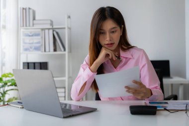 Genç Asyalı kadın masamda oturmuş elinde veri ve grafik kağıdıyla dizüstü bilgisayarını kullanıyor, hesapları analiz ediyor, iş geliştirme stratejileri planlıyor..