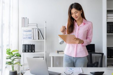 Genç Asyalı kadın masamda oturmuş elinde veri ve grafik kağıdıyla dizüstü bilgisayarını kullanıyor, hesapları analiz ediyor, iş geliştirme stratejileri planlıyor..