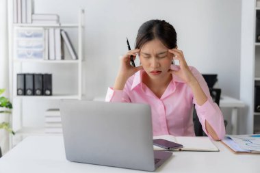 Stres altında çalışan genç Asyalı kadın işçi başını kaşıyormuş gibi yapıyor çünkü masadaki dizüstü bilgisayar ve belgelerle çalışmaktan çok yorulmuş..