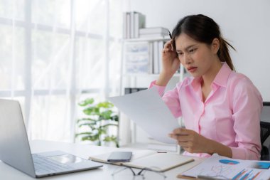 Stres altında çalışan genç Asyalı kadın işçi başını kaşıyormuş gibi yapıyor çünkü masadaki dizüstü bilgisayar ve belgelerle çalışmaktan çok yorulmuş..