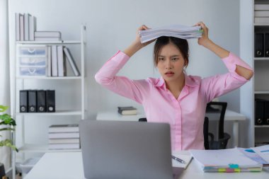 Stres altında çalışan genç Asyalı kadın işçi başını kaşıyormuş gibi yapıyor çünkü masadaki dizüstü bilgisayar ve belgelerle çalışmaktan çok yorulmuş..