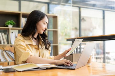 Evden çalışan hırslı Asyalı kız dizüstü bilgisayar ekranlarına ve mali belgelere bakıyor, kadın uzaktan çalışırken e-postaları kontrol ediyor veya araştırma yapıyor.