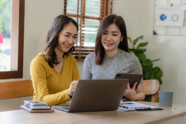 Dizüstü bilgisayarlar ve finansal belgelerle çalışan iki Asyalı kadın pazarlama planlarını tartışıyor. Yeni şirketin satış stratejisi güncel olaylarla uyumlu.