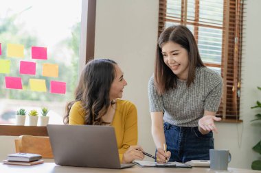 Dizüstü bilgisayarlar ve finansal belgelerle çalışan iki Asyalı kadın pazarlama planlarını tartışıyor. Yeni şirketin satış stratejisi güncel olaylarla uyumlu.