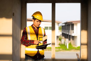 Genç Asyalı inşaat mühendisi ya da miğfer takan inşaat yöneticisi inşaat alanını incelerken yüzünü çeviriyor ve gülümsüyor..
