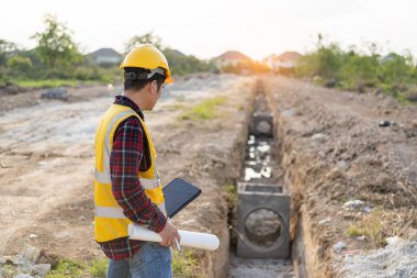 Genç Asyalı inşaat mühendisi ya da miğfer takan inşaat yöneticisi inşaat alanını incelerken yüzünü çeviriyor ve gülümsüyor..