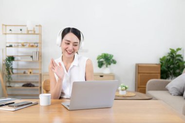 Kulaklık takan gülümseyen Asyalı kız bilgisayarını kullanarak ekranda çalışırken müzik dinlemekten mutlu oluyor. Öğrenci dili öğrenme ve ev konseptinde çalışma konusunda heyecanlı