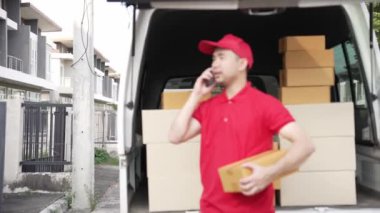 Elinde karton bir kutu tutan yakışıklı bir Asyalı arka planda minibüs olan bir evin önünde duruyor. Teslimat konsepti. 4K çözünürlüğü.