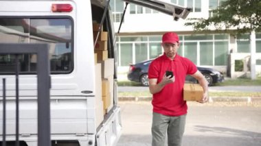 Elinde karton bir kutu tutan yakışıklı bir Asyalı arka planda minibüs olan bir evin önünde duruyor. Teslimat konsepti. 4K çözünürlüğü.