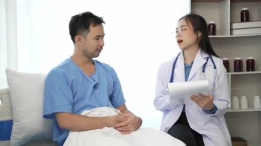 Genç Asyalı kadın doktor ve genç erkek hasta nabzını kontrol ederken yatıyorlar. Danışmak ve hemşirelerin not alıp Video 4k 'ye yardım etmelerini sağlamak.