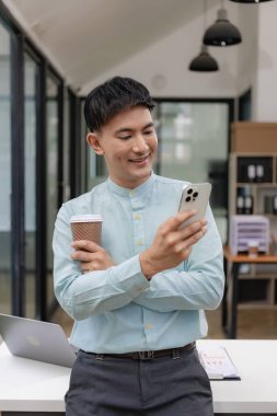 Genç Asyalı işadamı ofisinde ve kollarını kavuşturup gülümsüyor. Genç Asyalı iş adamı, başarılı profesyonel, milenyum lideri kollarını bilgisayar ekranının önünde birleştirerek şirket ofisinde duruyor..