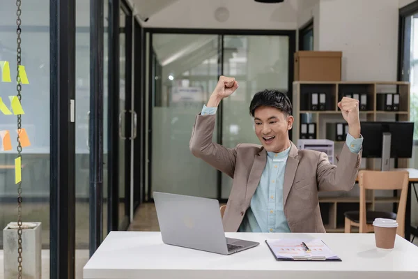 A businessman wearing a formal suit is at work. He sat in front of his laptop computer, raised his hand and shouted with great joy. About success in business and celebrations