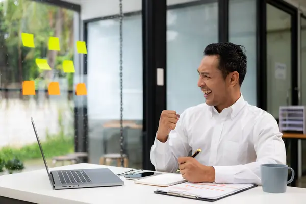 A businessman wearing a formal suit is at work. He sat in front of his laptop computer, raised his hand and shouted with great joy. About success in business and celebrations