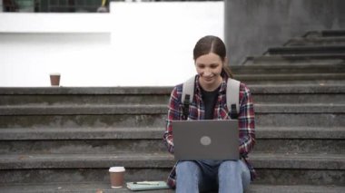 Güzel sarışın kız öğrenci kampüsün dışındaki merdivenlerde oturuyor. Üniversite eğitimi, e-öğrenme, dizüstü bilgisayar, öğrenme.