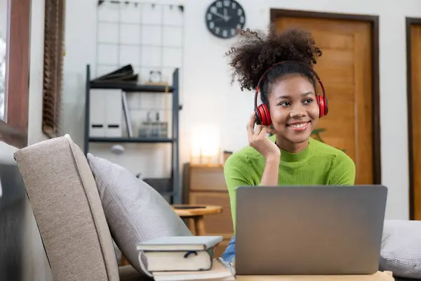 African American girl in headphones watching video lesson on computer in living room at home, happy in headphones having online class using laptop