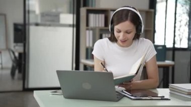 Z jenerasyonundaki kulaklıklı genç kız laptopta internet okuyor. Webinars 'ın sesli anlatımını dinliyor. Notlar yazıyor, podcast dinliyor, dinleme dersleri alıyor.