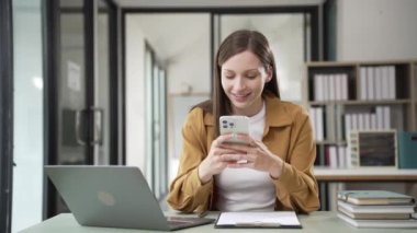 Uzak kariyer. Cep telefonuyla konuşan ve evde dizüstü bilgisayar kullanan gülümseyen bir kadın. Ev bilgisayarında çalışırken cep telefonu kullanan genç serbest çalışan bir kadın. Yüksek kalite 4k görüntü