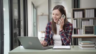 Uzak kariyer. Cep telefonuyla konuşan ve evde dizüstü bilgisayar kullanan gülümseyen bir kadın. Ev bilgisayarında çalışırken cep telefonu kullanan genç serbest çalışan bir kadın. Yüksek kalite 4k görüntü