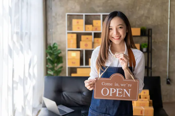Happy young business owner standing holding open sign Happy Asian woman wearing an apron hanging a shop opening sign smile happily Proud in personal business