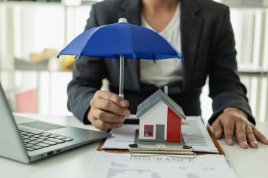 Ahşap bir masa üzerindeki ev modelini kapsayan mavi şemsiye emlak sigortası, ipotek sigortası konsepti emlak işinde risk kontrolüdür..