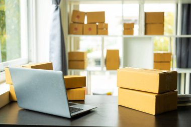 Raflardaki paket kutuları ve masadaki dizüstü bilgisayarın yanındaki renkli alışveriş torbaları. Evden online alışverişte KME işi.