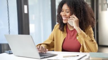 İş elbisesi içindeki güzel Afrikalı kadın ofiste çalışırken cep telefonuyla konuşuyor. Modern akıllı telefonu kullanarak 4K danışma için müşteriyle konuşuyor.
