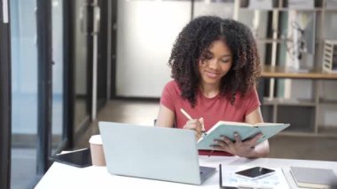 Diz üstü bilgisayar kullanan Afrikalı bir kız ödev için notlar yazıyor, ödev yazıyor, ders çalışıyor. İnternetten öğrenen öğrenciler, üniversite sınavlarına 4k 60p.