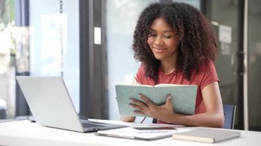 Diz üstü bilgisayar kullanan Afrikalı bir kız ödev için notlar yazıyor, ödev yazıyor, ders çalışıyor. İnternetten öğrenen öğrenciler, üniversite sınavlarına 4k 60p.