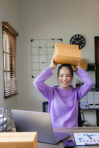 A purple-clad independent Asian woman SME uses a laptop and boxes to receive and review online orders to prepare packages to sell to customers. vertical picture