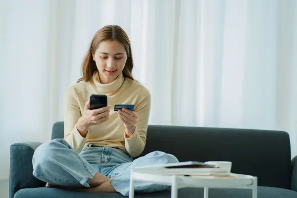 Young Asian woman sitting on sofa at home making online reservation on mobile phone using debit card to pay Woman doing online shopping using mobile device
