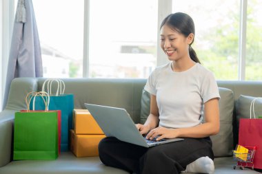 Akıllı telefon ve dizüstü bilgisayar kullanan neşeli Asyalı kadın, online alışveriş, kredi kartı, ödeme, evdeki koltukta oturmak, ödeme konsepti internet bankacılığı başvurusu yoluyla.