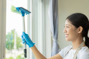 Güzel Asyalı ev hanımı ev işi yapıyor ve bir önlükle evi temizliyor, oturma odasının zeminini temizlemek için süpürge, paspas ve bez kullanıyor. Mutlu ev hanımı ev işi yapıyor.