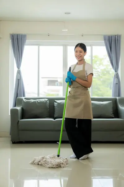 Beautiful Asian housewife doing housework and cleaning the house with an apron, using a vacuum cleaner, mop, cloth to clean the floor in the living room. Happy housewife doing housework