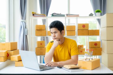 Genç bir Asyalı erkek KOBİ işletme sahibi ürünleri çevrimiçi satar ve çevrimiçi siparişler göndermek için ürün paketleme kutuları hazırlar ve çevrimiçi olarak alışveriş yapan müşterileri evde bulundurur..
