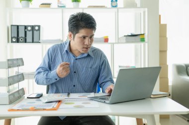 Home Man 'de çalışan yakışıklı Asyalı işadamı bilgisayarla iş yerinde performans analizi raporu hazırlayan dizüstü bilgisayarlı grafikleri ve çizelgeleri analiz ediyor..