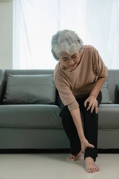 一位年迈的亚洲妇女因右脚踝疼痛而面带微笑 在沙发上坐下 走路走得太多而受了伤 — 图库照片