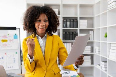 Masada oturmuş kameraya bakan neşeli bir iş kadını, finansal iş analizi için mali grafik belgesiyle çalışırken bilgisayarını kullanarak gülümseyen Afrikalı Amerikalı yönetici..