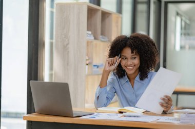 Masada oturmuş kameraya bakan neşeli bir iş kadını, finansal iş analizi için mali grafik belgesiyle çalışırken bilgisayarını kullanarak gülümseyen Afrikalı Amerikalı yönetici..