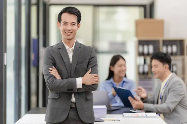 Asian businessman standing with arms crossed and looking at camera Standing in front of a table with co-workers who are meeting to discuss financial investments in the back of the office.