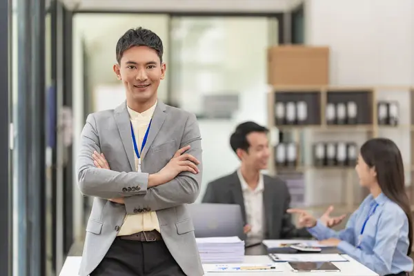 Asian businessman standing with arms crossed and looking at camera Standing in front of a table with co-workers who are meeting to discuss financial investments in the back of the office.