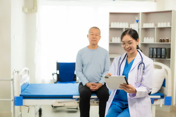 担当の看護師は 高齢者の世話をして 病院の部門の部屋で寝る アジアの女性医師は 彼の痛みと症状 生命保険の概念についてベッドで男性患者と話します — ストック写真