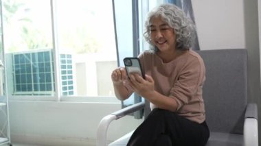 Kanepede oturan yaşlı kadın ya da Asyalı büyükanne internet üzerinden ödemek için cep telefonu kullanıyor, oyunlar oynuyor, film izliyor, müzik izliyor, sosyal medya evinde oturuyor..