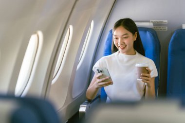 Genç Asyalı bir kadın uçakta oturuyor ve akıllı bir telefonla konuşuyor. Ekonomi sınıfında pencere kenarında otururken bir uçak uçuşu sırasında akıllı bir cep telefonu kullanıyor.