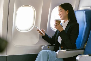 Genç Asyalı bir kadın uçakta oturuyor ve akıllı bir telefonla konuşuyor. Ekonomi sınıfında pencere kenarında otururken bir uçak uçuşu sırasında akıllı bir cep telefonu kullanıyor.