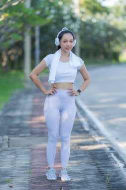 Beyaz spor giysili genç Asyalı kadın parkta koşup, dışarıda sağlıklı aktiviteler yapmadan önce kaslarını esnetiyor. Dikey resim.
