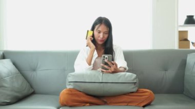Gülümseyen genç bir kadın cep telefonu kullanıyor, elektronik bankacılık başvurusu vasıtasıyla evdeki rahat bir koltukta ödeme yapıyor, online alışveriş yapıyor, internetten satın alıyor ve ödüyor..