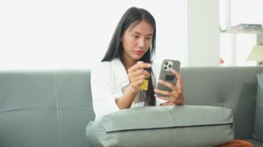 Gülümseyen genç bir kadın cep telefonu kullanıyor, elektronik bankacılık başvurusu vasıtasıyla evdeki rahat bir koltukta ödeme yapıyor, online alışveriş yapıyor, internetten satın alıyor ve ödüyor..