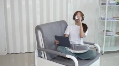 Asyalı kız rahat koltukta oturup tabletle mutlu bir şekilde oynuyor. Sabahı oturma odasındaki dijital tabletinde sosyal medyada gezinerek geçiriyor. Biraz zaman geçir..