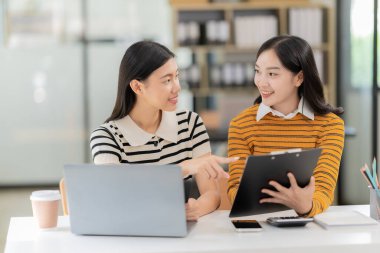 İki Asyalı kadın online satışlarda çalışarak gelirlerini kontrol ediyorlar. Satışlar istenen hedeflere uygun olduğu için dizüstü bilgisayarlarla.