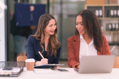 İki genç iş kadını laptoplarda çalışırken paneller hakkında konuşuyor, belgeler okuyor ve yeni bir projeyi tartışırken notlar yazıyorlar..
